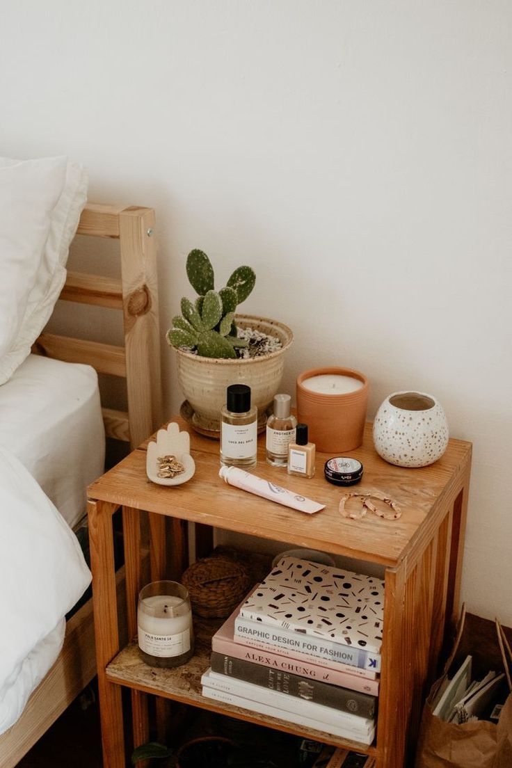 a wooden table topped with books and plants next to a white bed covered in pillows