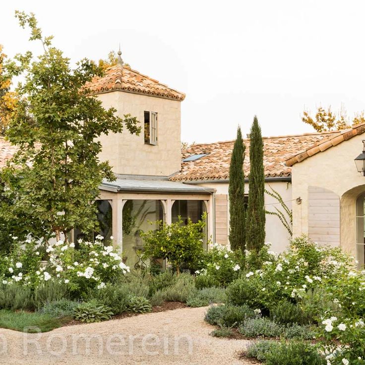 a house that is surrounded by trees and flowers in front of the house, with an entrance way leading to it