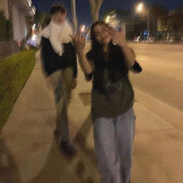 two people walking down a sidewalk at night with one person covering his eyes and the other holding her hand up