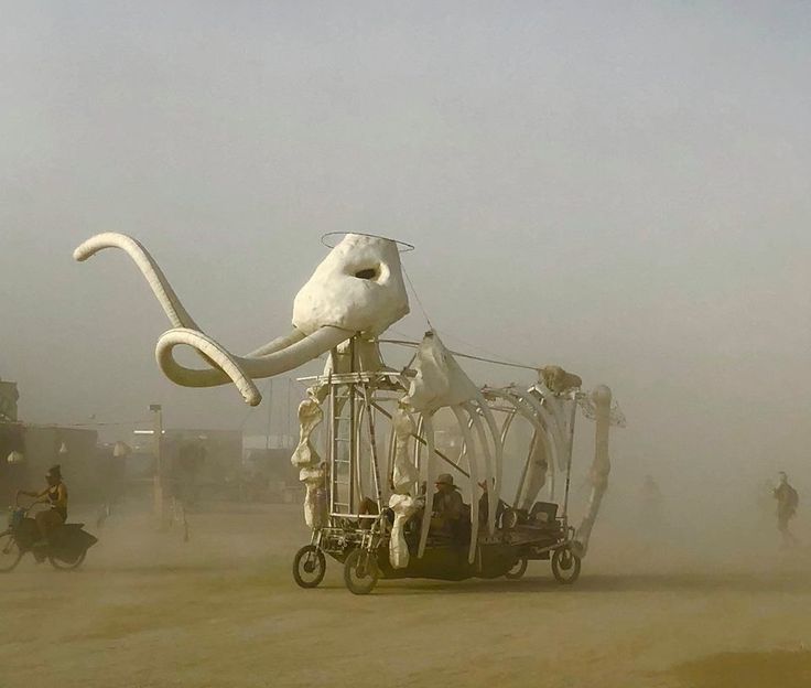 an octopus sculpture on wheels in the middle of a dirt field with people riding bikes behind it