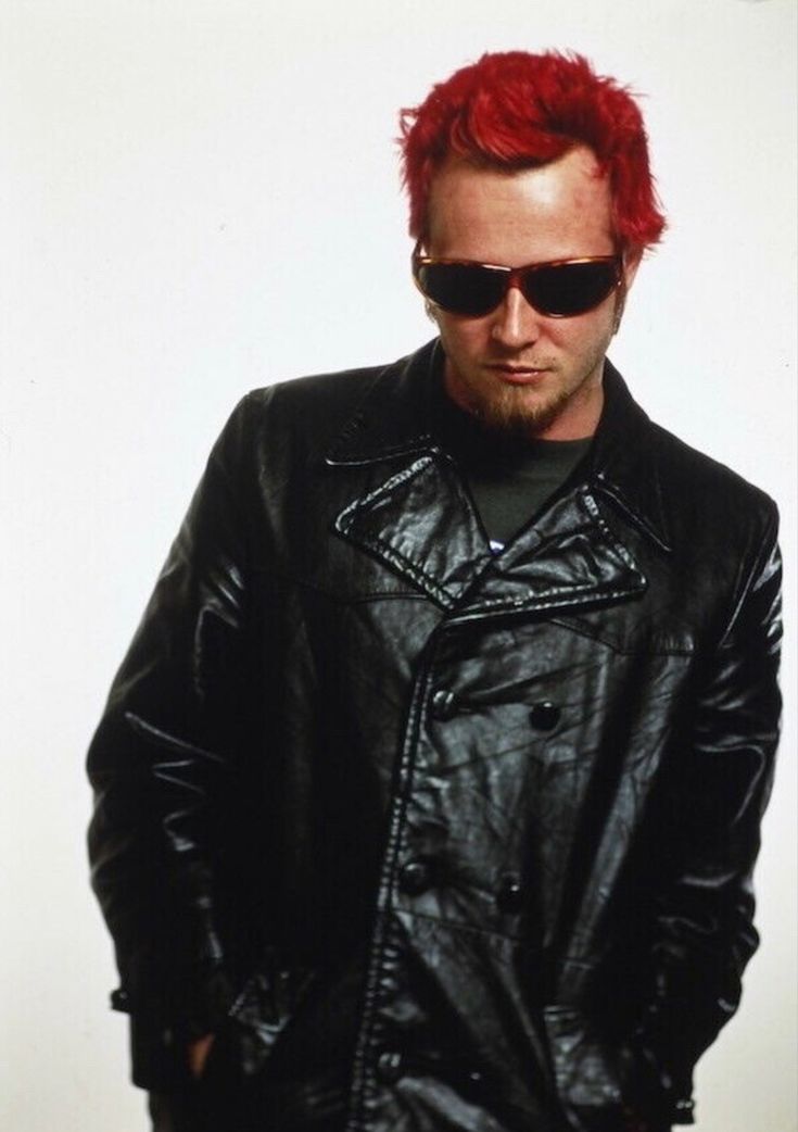 a man with red hair and sunglasses standing in front of a white wall wearing a black leather jacket