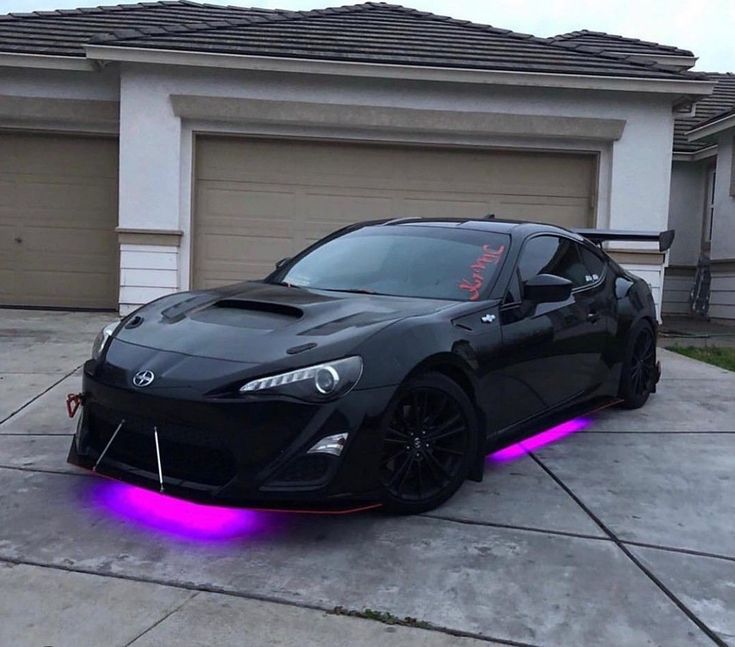 a black sports car parked in front of a house with purple lights on it's side