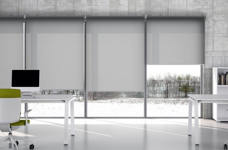 an office with two desks and chairs in front of a window that has roller shades on it