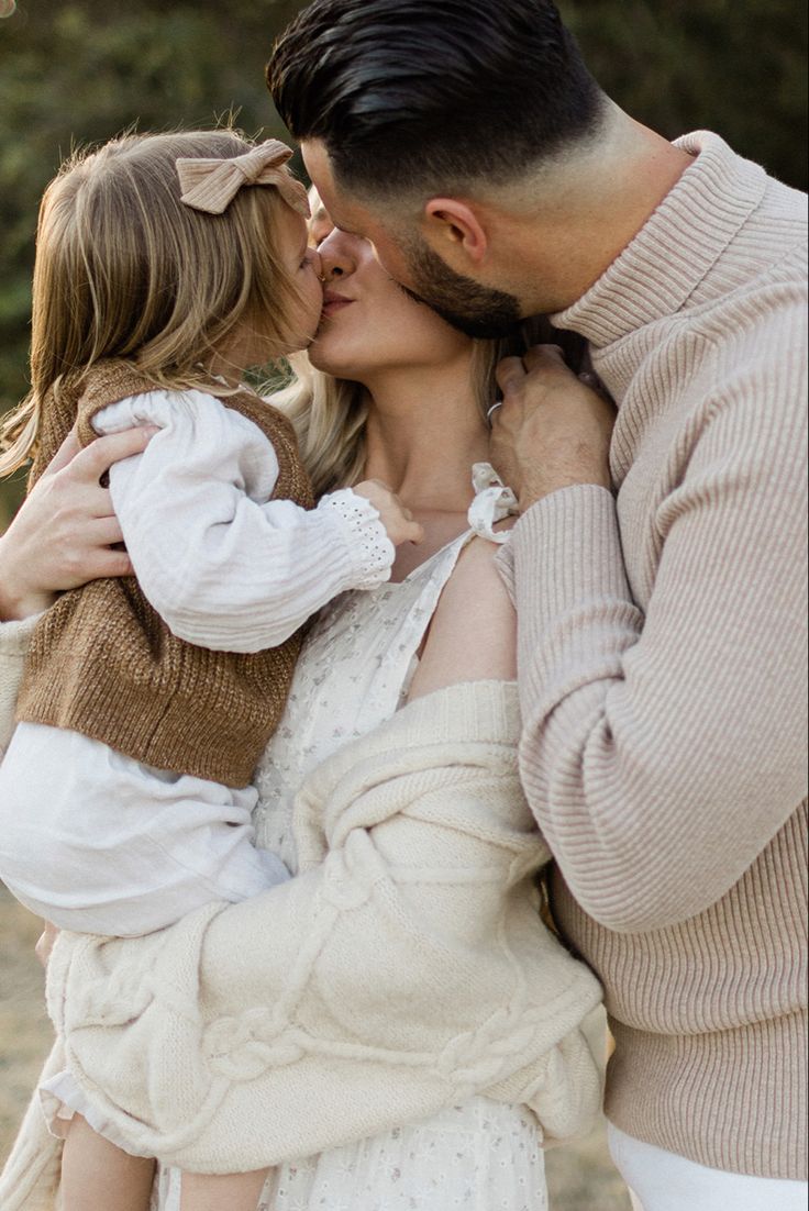 a man and woman kissing each other while holding a child in their arms, with trees in the background