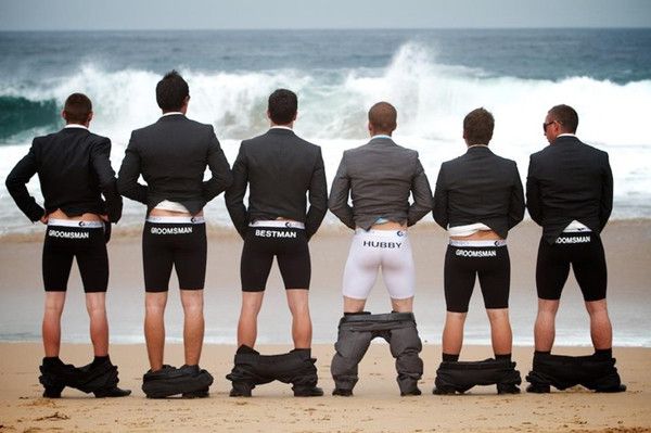 four men in black suits and white shorts standing on the beach with their backs to the camera