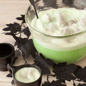 a green liquid in a glass bowl with leaves around it and two black cups next to it