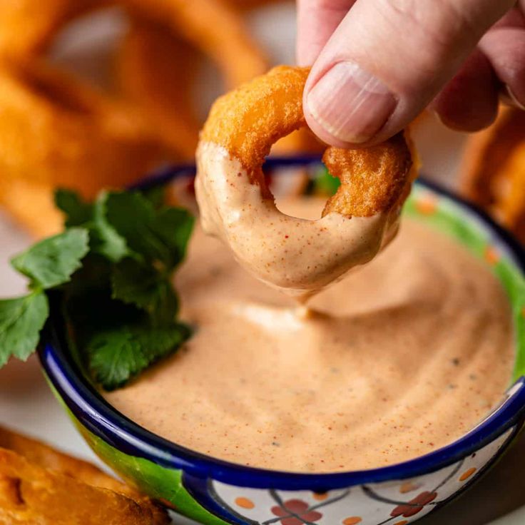 a person dipping sauce into a bowl with onion rings and cilantro on the side