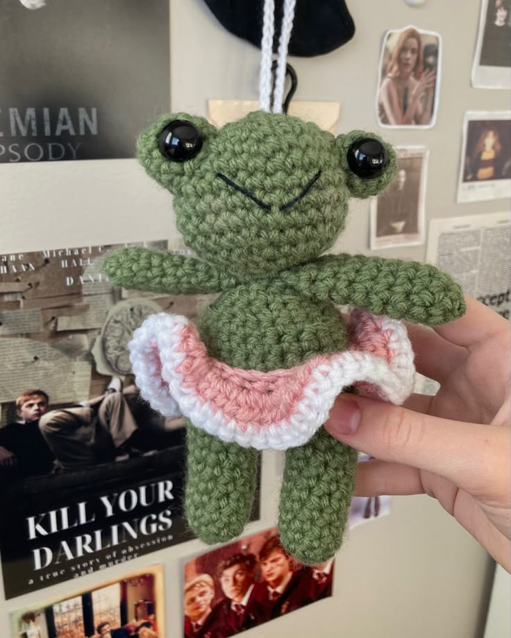 a crocheted frog doll hanging from a hook on a wall with posters in the background