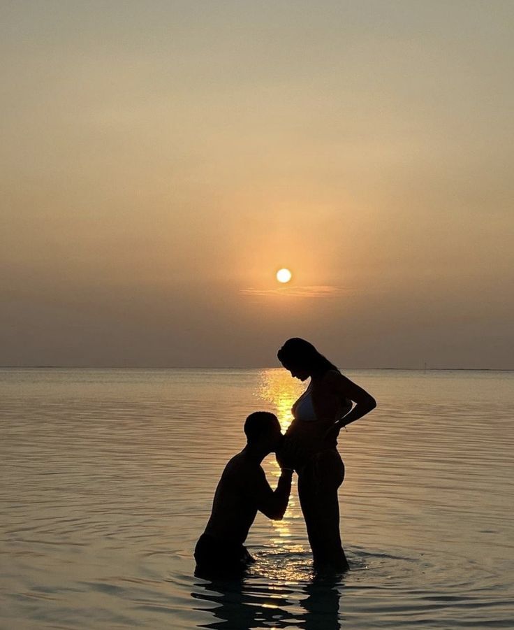 two people are standing in the water at sunset, one is touching the other's chest