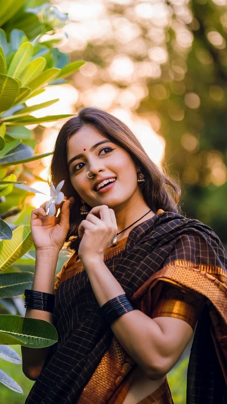 a beautiful woman standing next to a tree with a flower in her hand and smiling at the camera