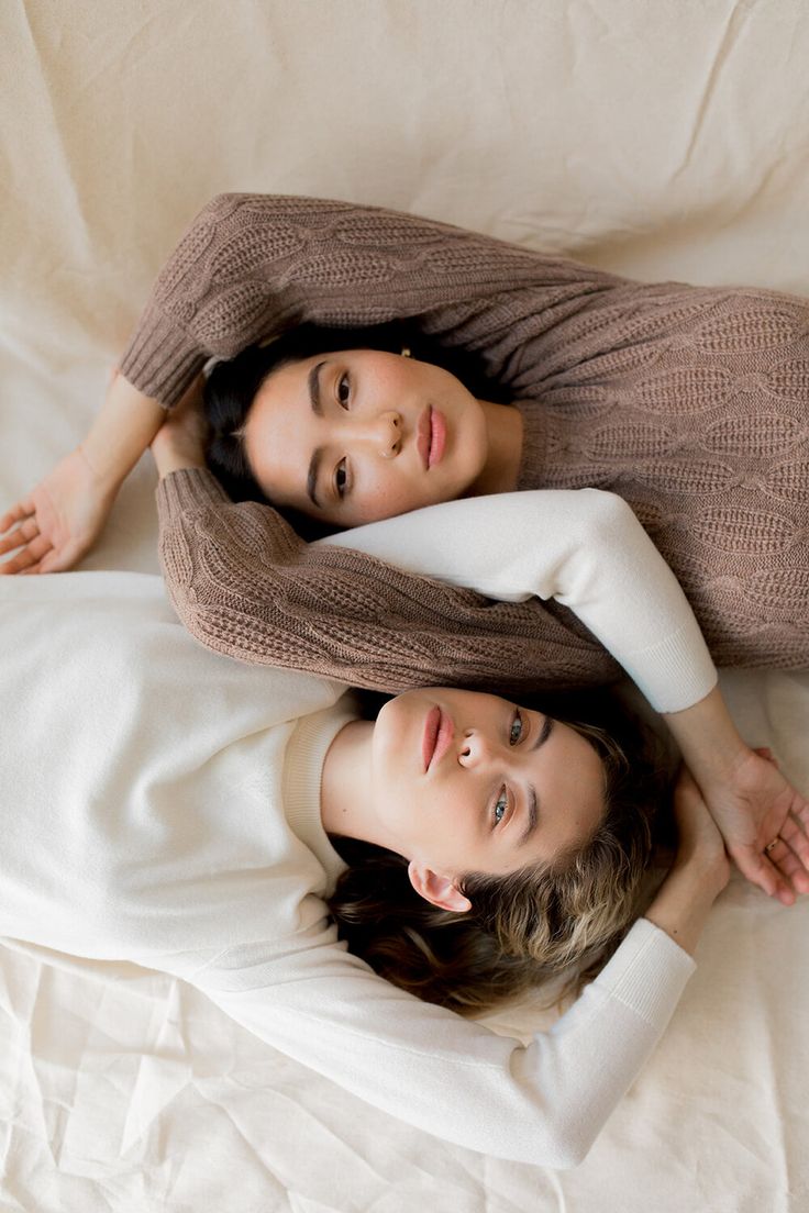 two women laying on top of each other on a white sheet covered bed with their arms around each other