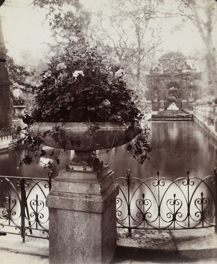 an old photo of a fountain with flowers in the center and trees around it, surrounded by wrought iron fencing