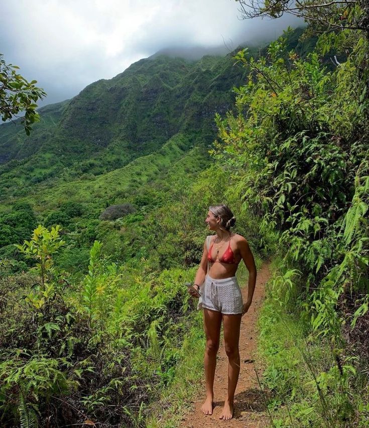 a woman standing on top of a dirt road next to a lush green forest covered hillside