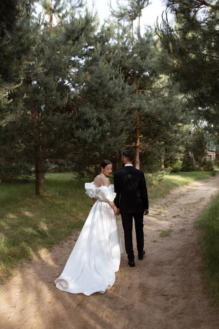 a bride and groom walking down a dirt path in the woods holding each other's hands