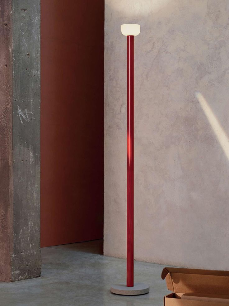 a red floor lamp sitting next to a box on the ground in front of a wall