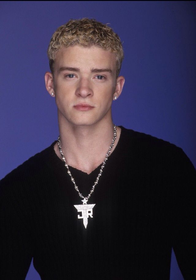 a young man with blonde hair wearing a black sweater and silver necklace, against a blue background