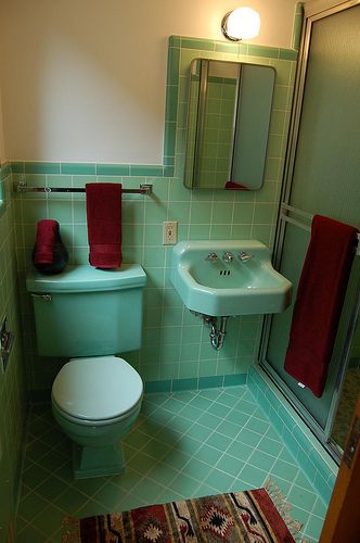 a bathroom with green tiles and a white toilet next to a sink, mirror and rug