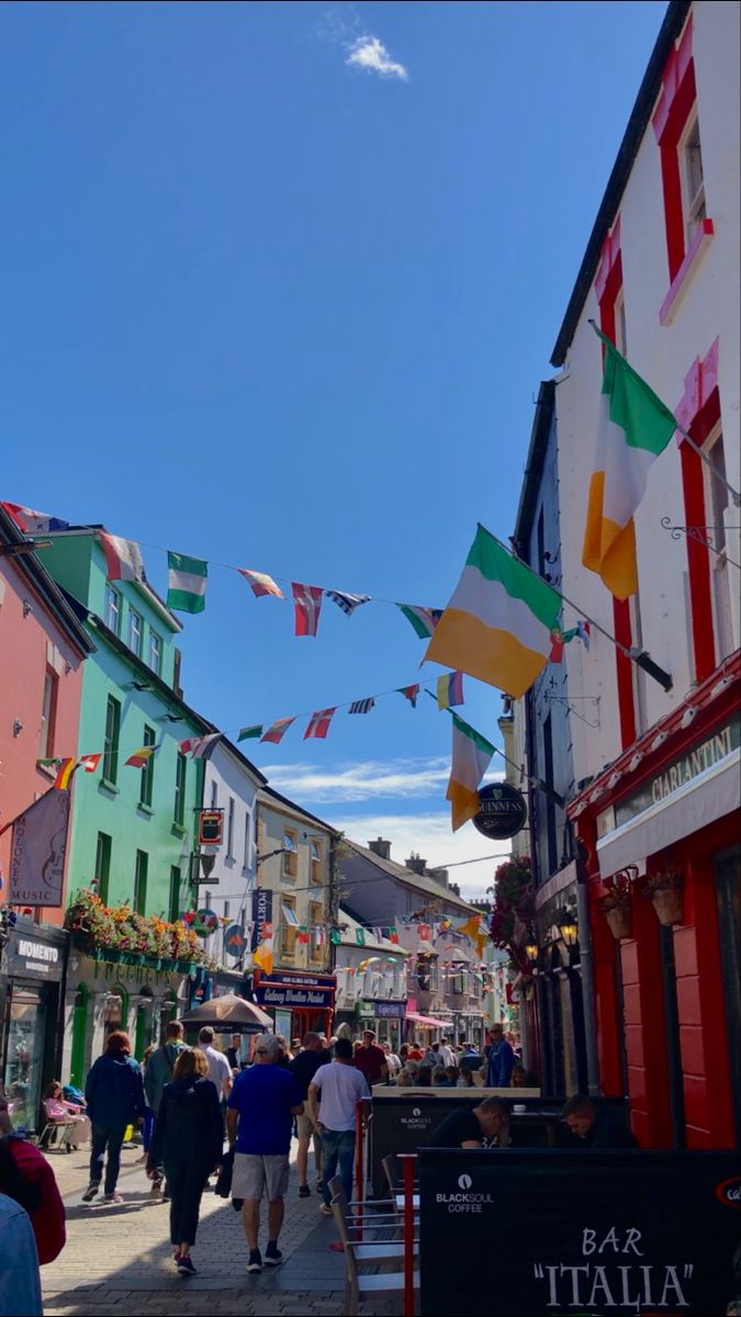 many people are walking down the street in front of some colorful buildings with flags flying above them