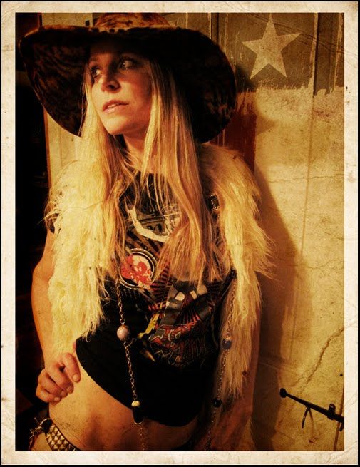 a woman with long blonde hair wearing a cowboy hat and feathered vest, standing in front of a wall