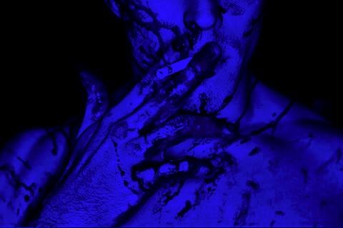 a man covered in blue paint with his hands on his face and chest, looking at the camera