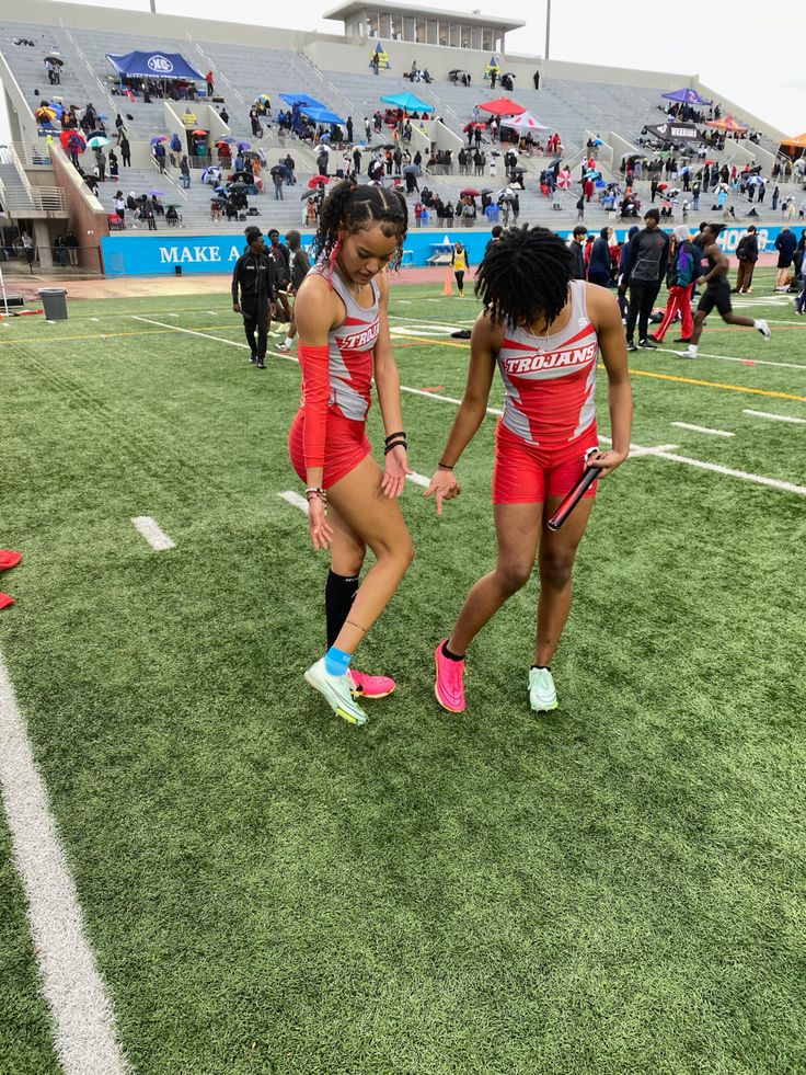 two girls in red and black uniforms are on the field at a football game with people watching