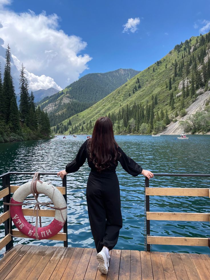 a woman standing on a dock next to a body of water with mountains in the background