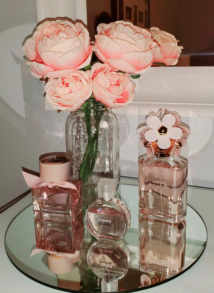 a glass table topped with pink flowers and perfume bottles next to each other on top of a white wall