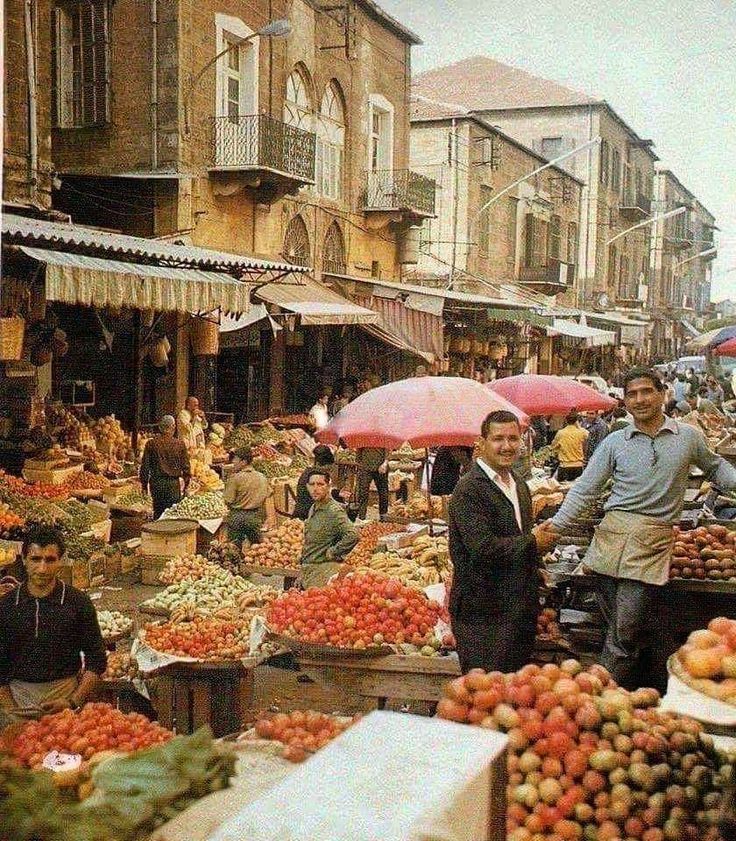 an outdoor market with lots of produce and people standing around it in front of buildings