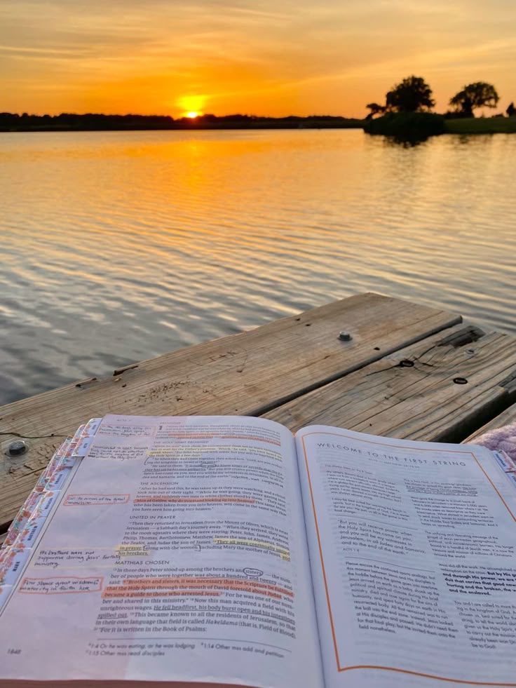 an open book sitting on top of a wooden dock next to the water at sunset