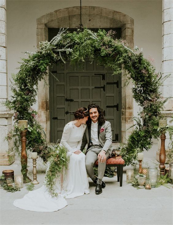 a bride and groom sitting on a bench in front of an arch with greenery