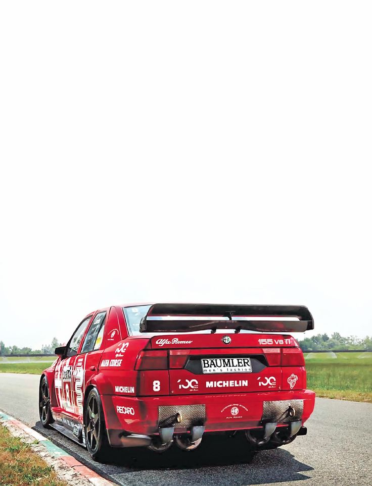 a red car driving down a road next to a grass covered field on a cloudy day