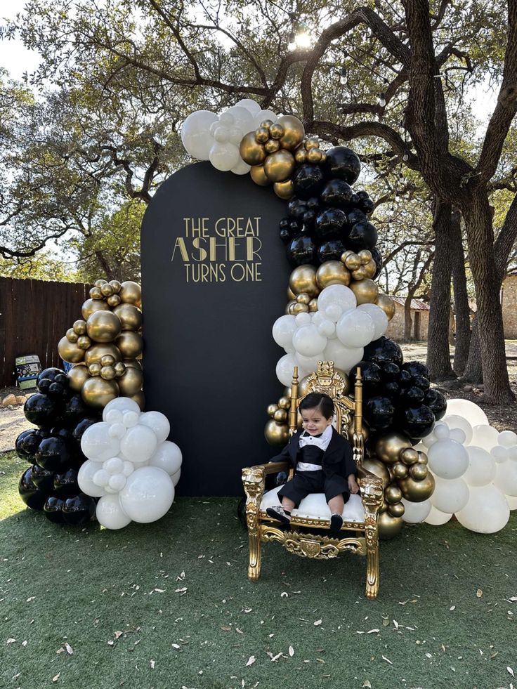 a little boy sitting on a chair in front of a giant balloon arch with black and gold balloons