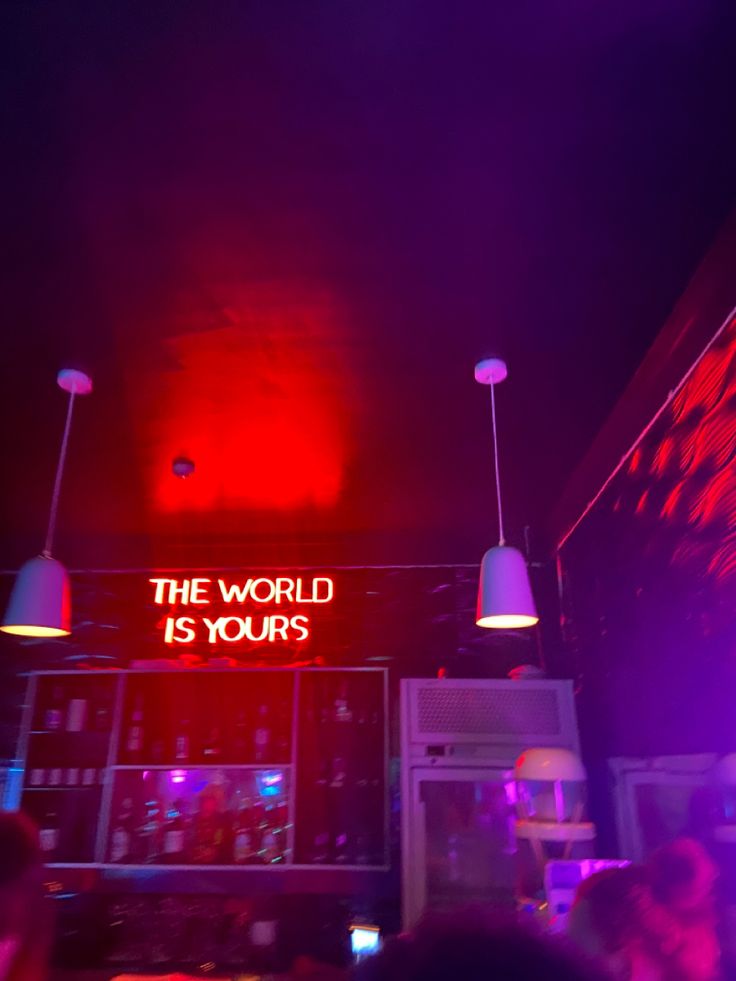 the world is yours sign lit up in purple and red lights above a bar with people sitting at tables