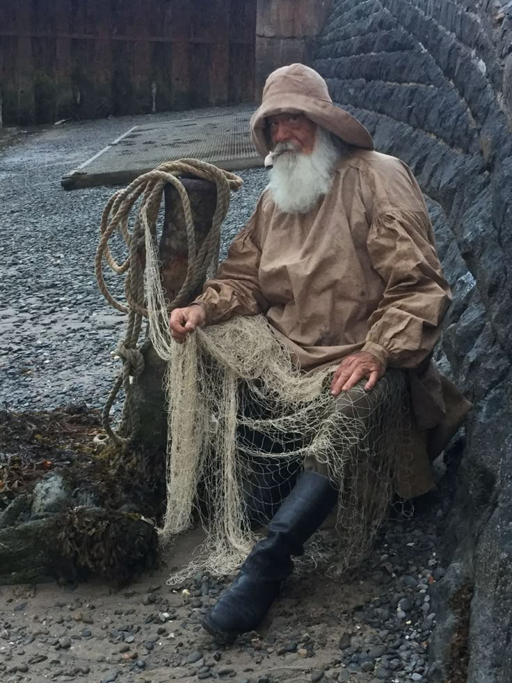 an old man sitting on the ground next to a stone wall wearing a hat and holding a fishing net