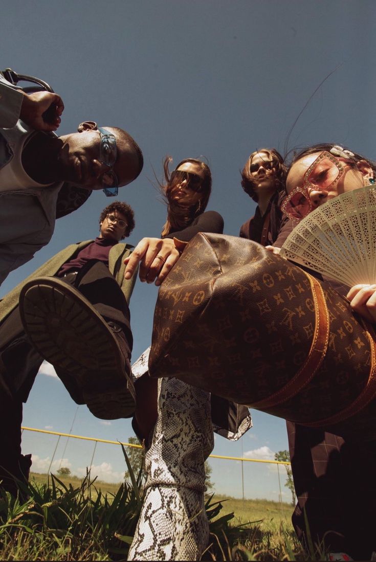 four people standing in the grass with one person holding an umbrella and another man wearing sunglasses