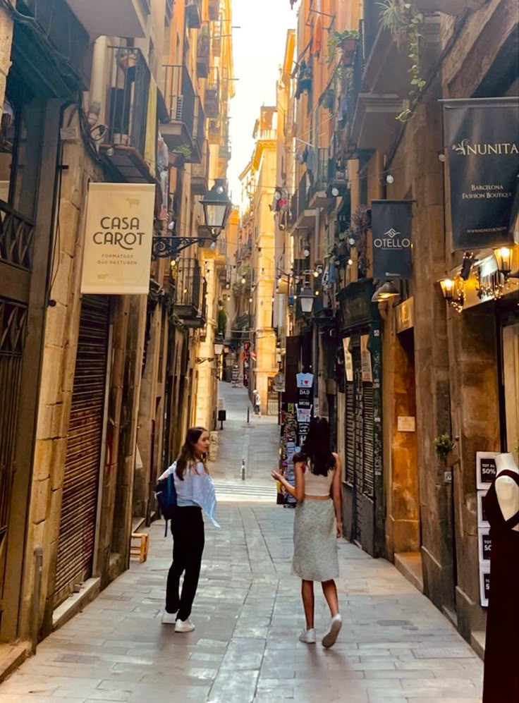 two women walking down an alley way in the middle of town with shops on both sides