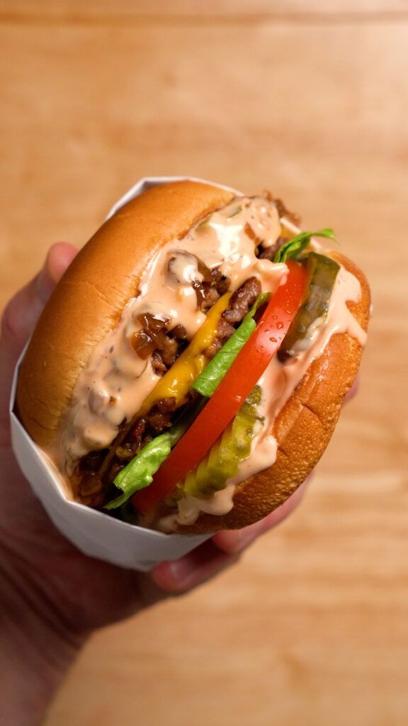 a hand holding a sandwich with meat, cheese and vegetables in it on a wooden table