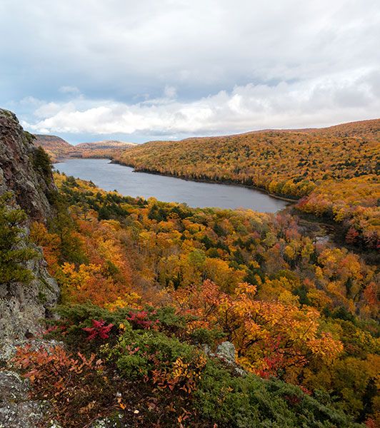 a lake surrounded by trees with fall colors in the foreground and cloudy skies above