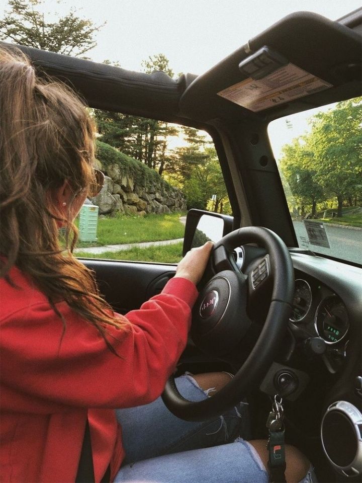 a woman driving a car down a street next to a lush green park on a sunny day