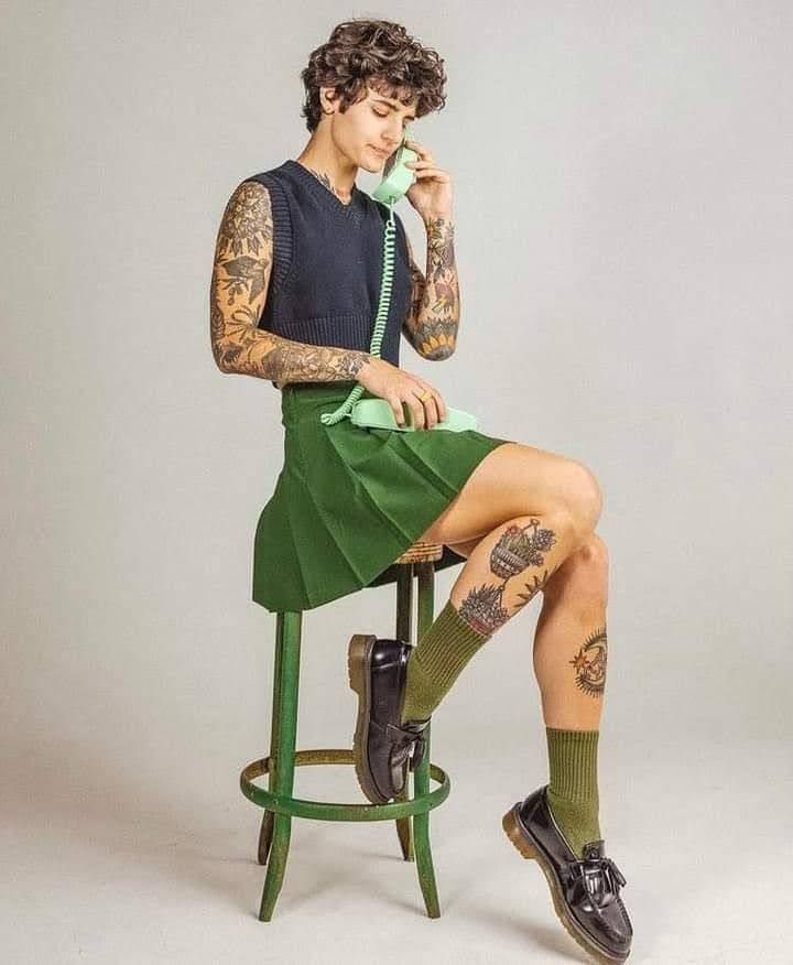 a man with tattoos sitting on a stool talking on a phone while holding a cell phone to his ear