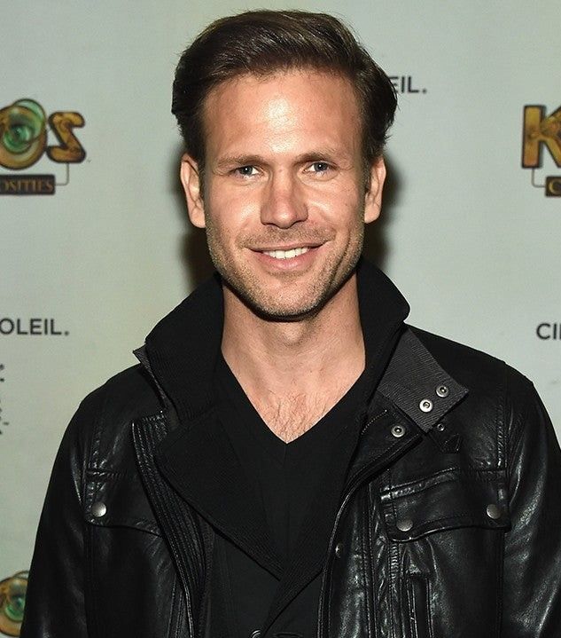 a man wearing a leather jacket and black shirt smiling at the camera while standing in front of a wall