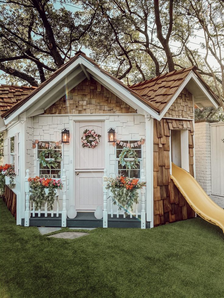 a small white house with flowers and a slide in the grass next to some trees