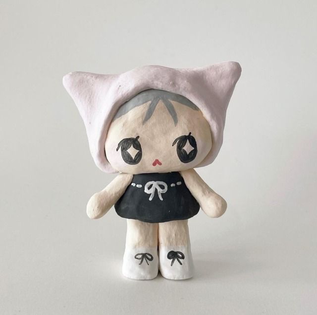 a small doll with black and white hair wearing a pink hat, sitting on a white surface