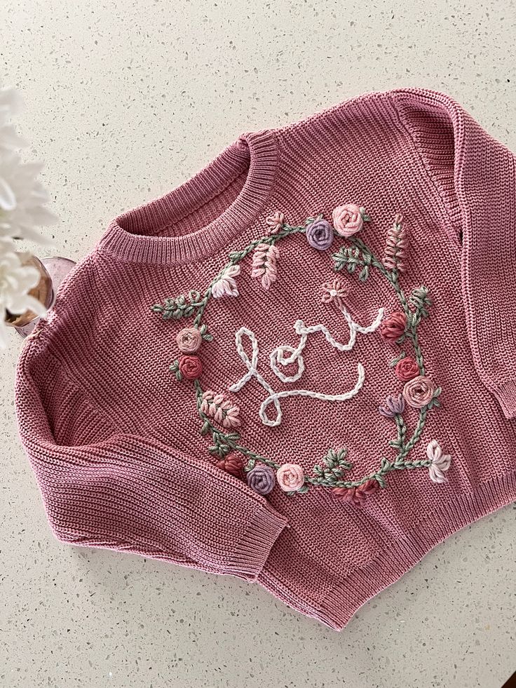 a pink sweater with the word love written on it and flowers in front of it