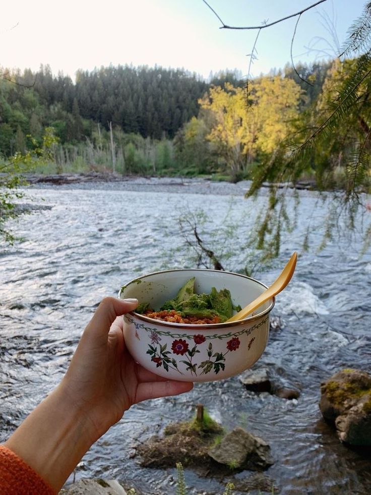 a person holding a bowl with food in it near a river and trees on the other side