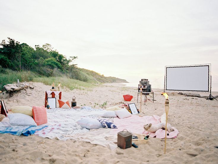 an outdoor movie set up on the beach with blankets, pillows and other items laid out