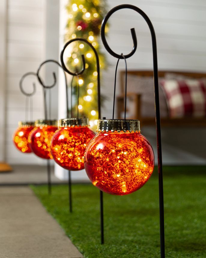 three red glass balls are hanging from black metal stands on the grass near a christmas tree