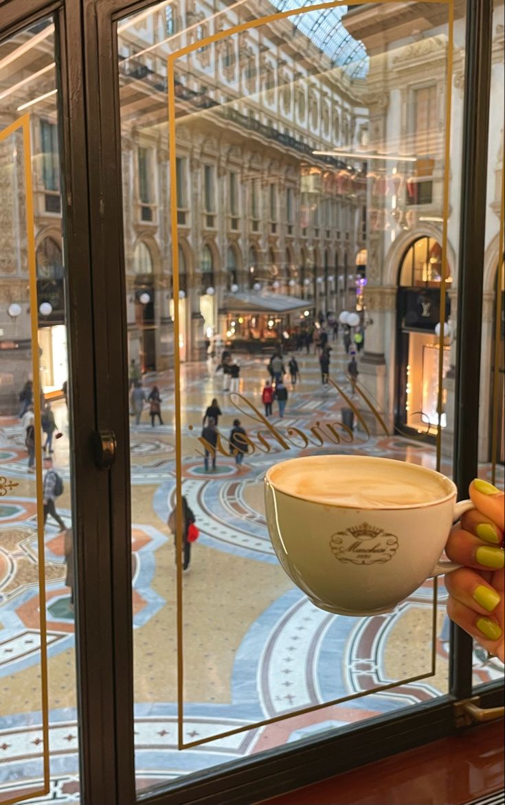 a person holding a cup of coffee in front of a window with people walking around