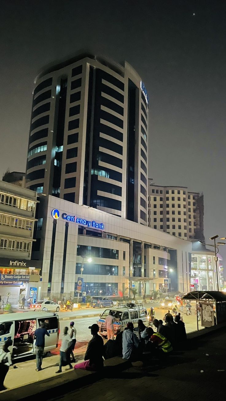 people are sitting on the curb in front of a large building at night with cars parked nearby