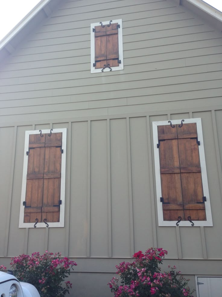 two windows with wooden shutters on the side of a gray building next to pink flowers